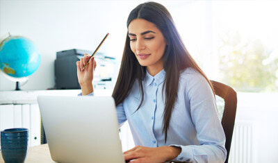 A woman with long dark hair is seated at a desk, working on a laptop. She holds a pencil in one hand and has a cup on the desk. A globe and a printer are visible in the background, as she reviews information related to BASE® HRA.