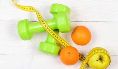 Two green dumbbells, a yellow measuring tape, two oranges, and a green apple on a white wooden surface—essential tools for staying true to your healthy resolutions.