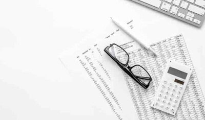 A calculator, a pair of glasses, and financial documents with numbers related to taxes on a white table, next to a keyboard.