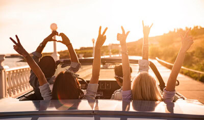 Four people in a convertible car driving on a road during sunset, with their arms raised in various celebratory gestures—it's the perfect image of Spring Break adventure.