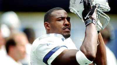 Football player in a white and blue jersey, resembling Darren Woodson, holding a helmet above his head, looking off into the distance. Other players and background details are blurred.