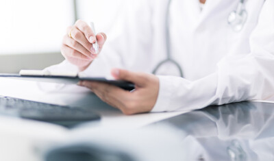 A physician in a white coat holding a pen and clipboard, seated at a desk with a computer keyboard in the foreground, ready to prescribe electronically.