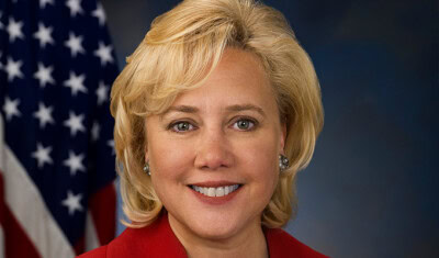 A smiling woman with short blond hair in a red jacket poses in front of an American flag, representing the spirit of innovation and small business.