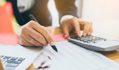 A person is using a pen to check off items on a printed list while operating a calculator, sitting at a desk with a laptop nearby, diligently organizing tips for new business strategies.