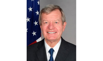 Portrait of a smiling man in a suit and tie, standing in front of an American flag, embodying the spirit of business and creating jobs.
