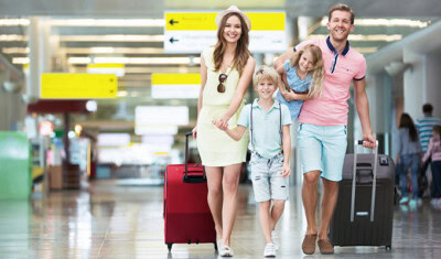 A family of four walks through an airport, each carrying or pulling luggage. Signs in the background point to various directions, hinting at their excitement for traveling abroad. The parents and children are smiling wide, clearly enjoying their journey together.