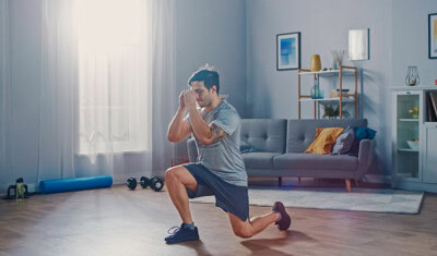 A man in a gray t-shirt and shorts exercises in a living room, performing a lunge with hands held close to his face. Exercise equipment is scattered around the room, showcasing the family benefits of staying active together.