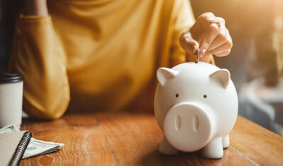 In uncertain times, a person wearing a yellow shirt places a coin in a white piggy bank on a wooden table, with dollar bills and a coffee cup nearby, showcasing ways to save and stretch your dollar.