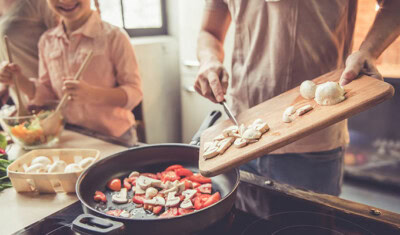 A person is adding sliced mushrooms from a wooden cutting board into a frying pan with chopped vegetables while another person in the background, after their Telemedicine appointment, is holding chopsticks near a salad.