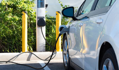 A white electric car is plugged into a charging station in a parking lot, with greenery visible in the background, highlighting the seamless blend of nature and technology. In case of any issues, roadside assistance for electric cars ensures peace of mind during your eco-friendly journey.