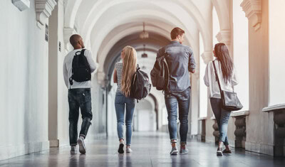 Four students with backpacks walk down a long, arched hallway, their backs turned to the camera. The hallway features ornate columns and tiled floors. They seem deep in discussion, perhaps about the Alliance Scholarship deadline approaching soon.