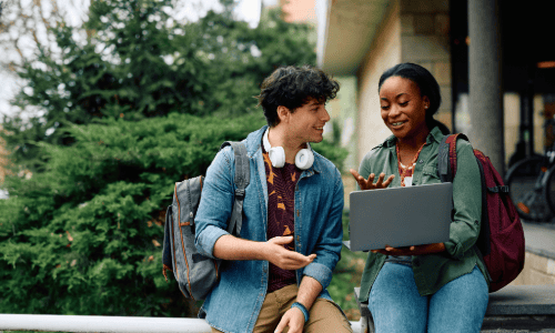Two students with backpacks sit outside, talking and looking at a laptop together. As they discuss, both seem engaged and focused on college student safety while sharing tips for a positive campus experience.