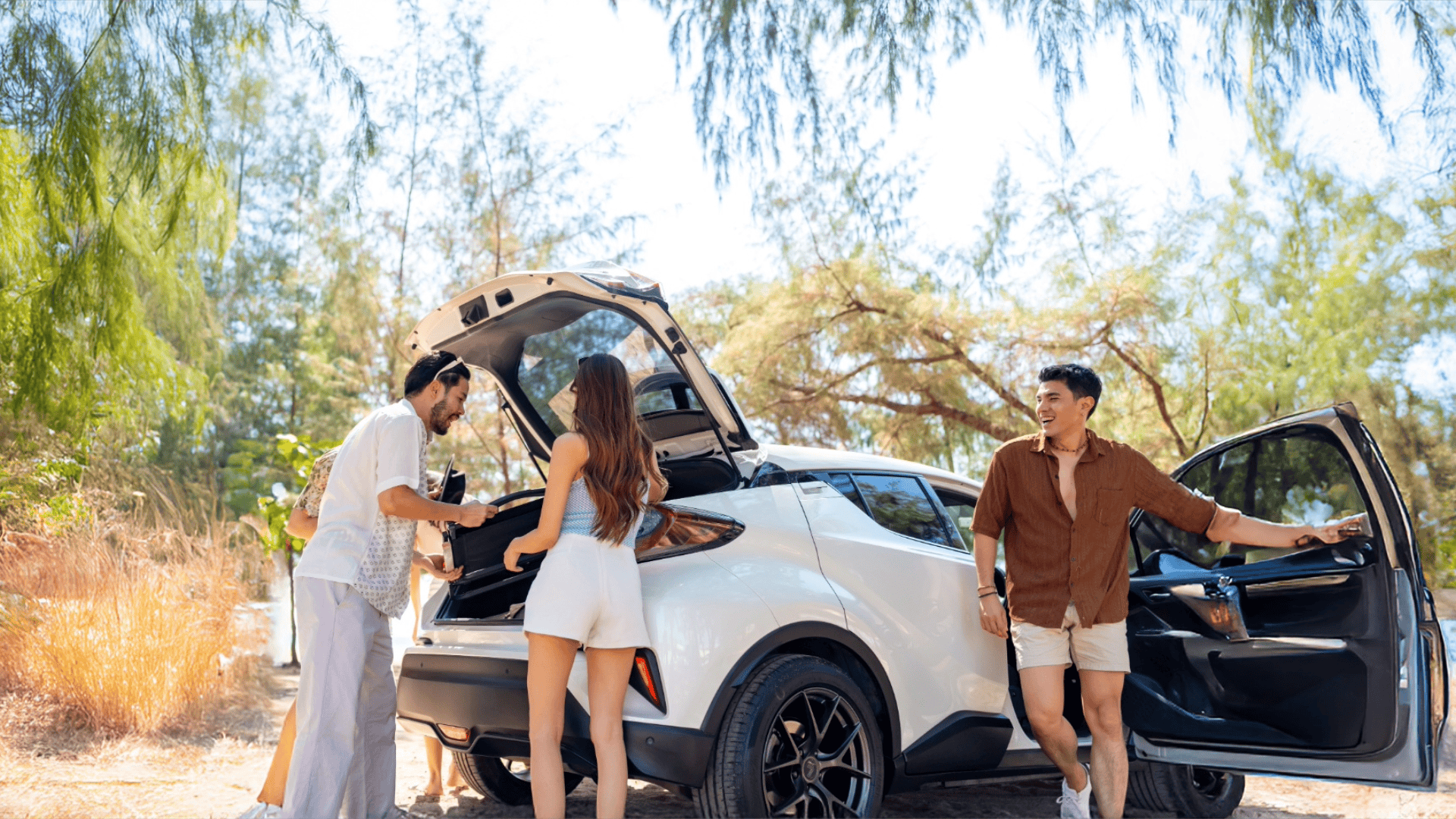 Four people stand beside a white SUV with the trunk open, preparing for an outdoor activity on a sunny day. Trees and dry grass are visible in the background, as they discuss ways to save money on travel with exclusive Alliance Travel Membership perks.