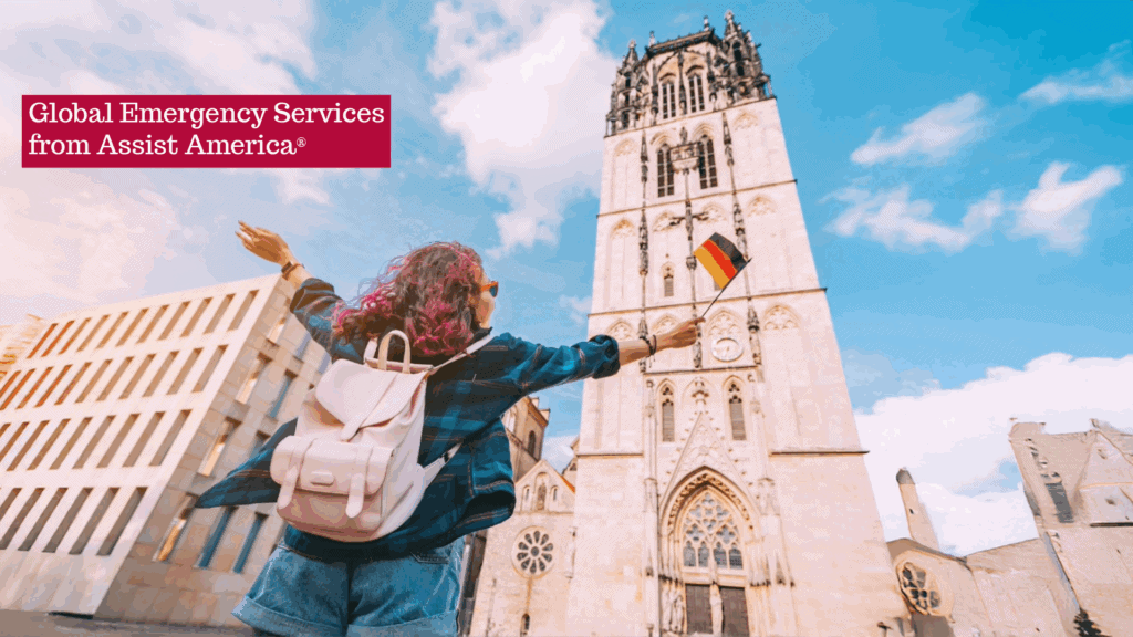 A person with a pink backpack stands in front of a tall historic tower, holding a small German flag; text reads "Global Emergency Services and travel protection from Assist America.