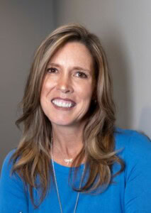 Rebecca Jackson, with long, light brown hair, wears a blue top and necklace, smiling warmly at the camera against a plain background.