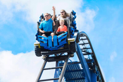 Three people, two adults and one child, ride a blue roller coaster on a bright, sunny day. The adults appear enthusiastic, with one raising their hand, while the child looks apprehensive. It's the perfect blend of entertainment for families seeking thrilling travel experiences with exciting discounts.