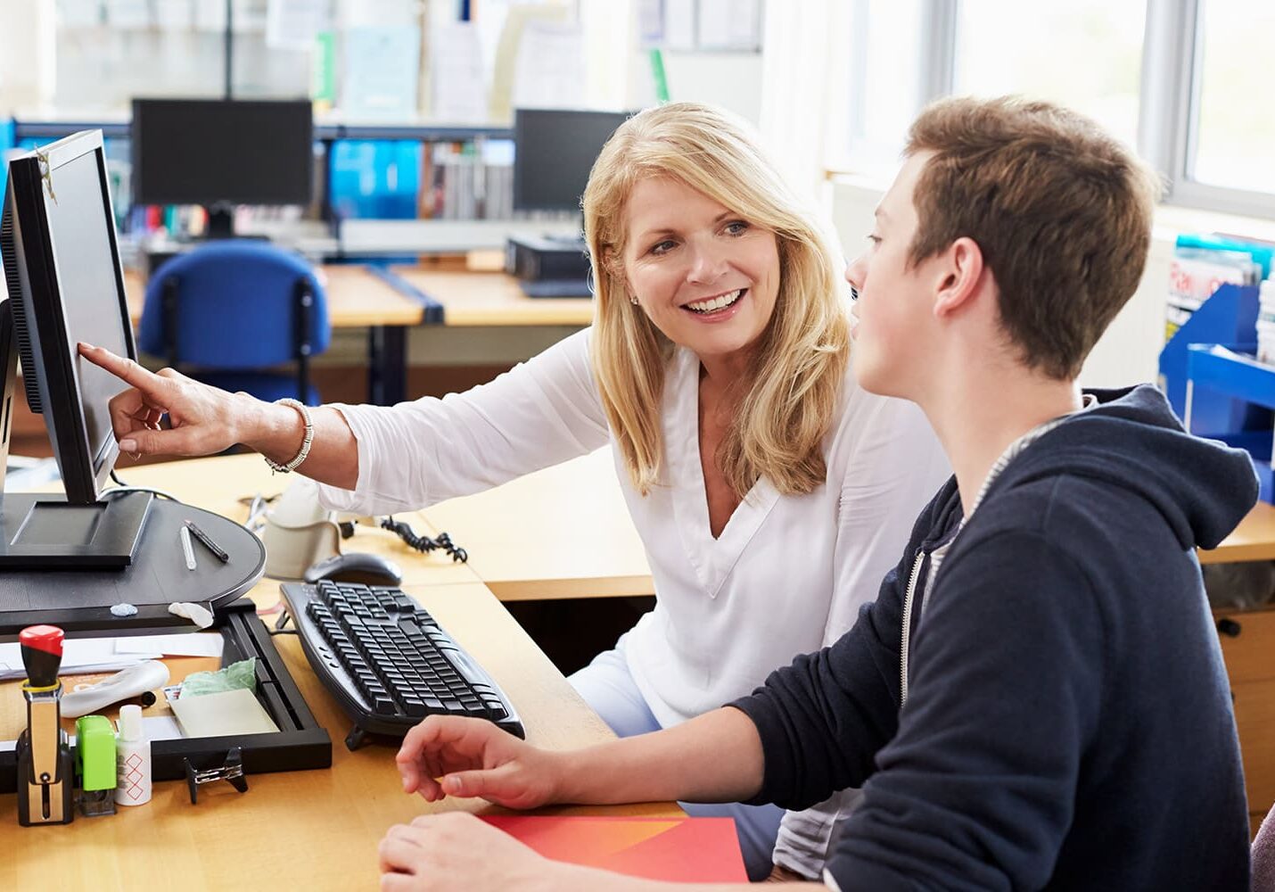 A woman points at a computer screen while talking to a young man sitting beside her in an office with desks and computers, discussing the best time to apply to college.
