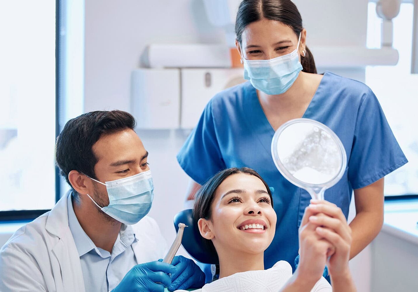 A dentist and dental assistant, both wearing masks, attend to a smiling patient in a dental chair who is holding up a mirror and looking at her teeth. The trio forms a cheerful group focused on maintaining dental health.