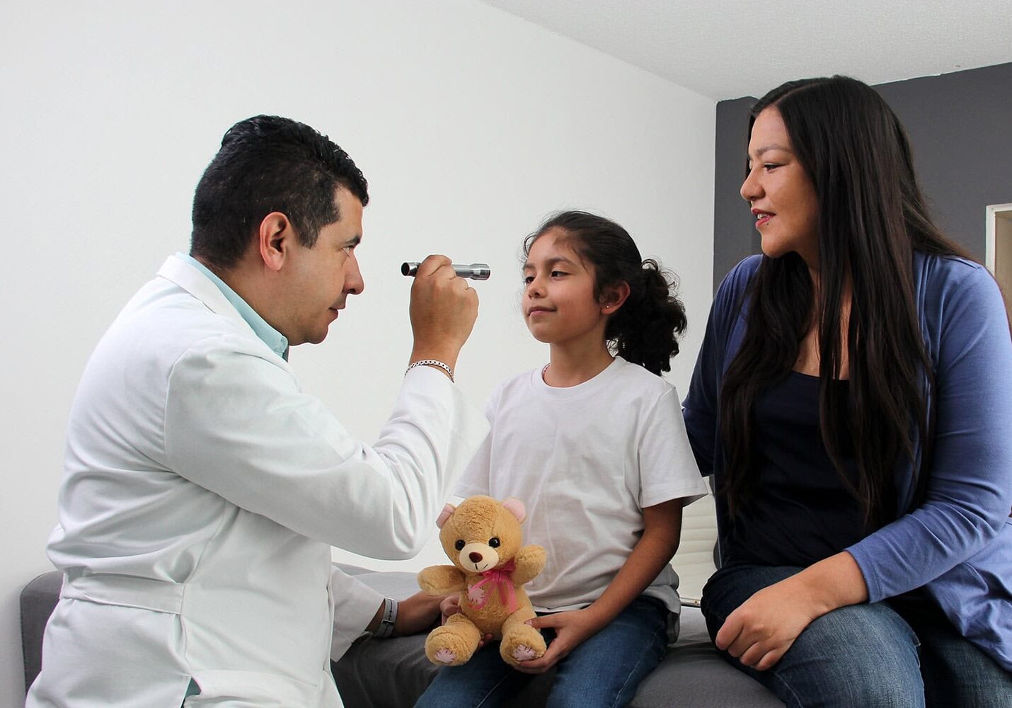 eye-doctor A doctor examines a young girl using an otoscope while she holds a teddy bear, illustrating the direct benefits of the healthcare alliance. A woman sits beside them, observing the interaction.
