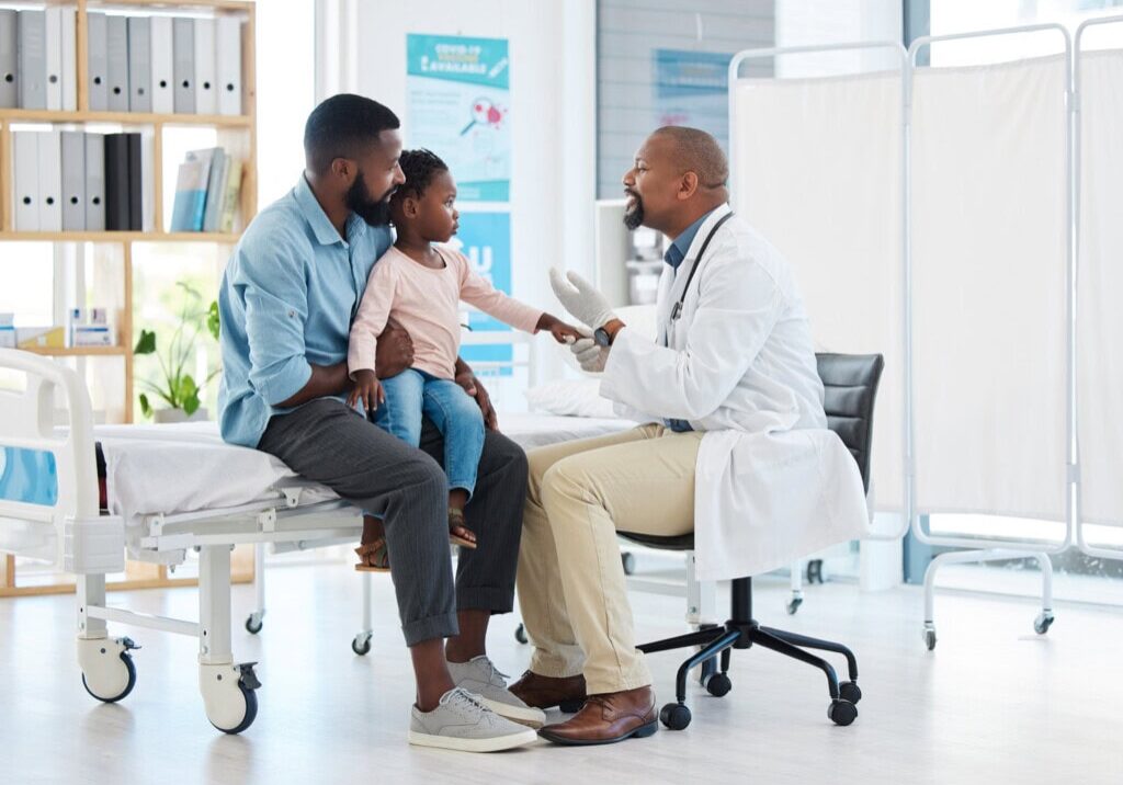 A doctor sits on a chair, engaging with a young girl sitting on her father's lap in a medical examination room, fostering an atmosphere where families feel cared for and supported.