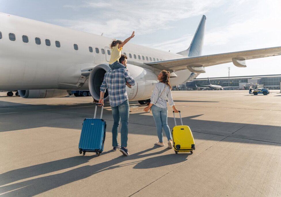 A family with two adults and one child walks towards an airplane on the tarmac, each adult pulling a suitcase. The child sits on one adult’s shoulders, pointing towards the aircraft. This charming moment highlights the joy of family activities and strengthens their family relationships.