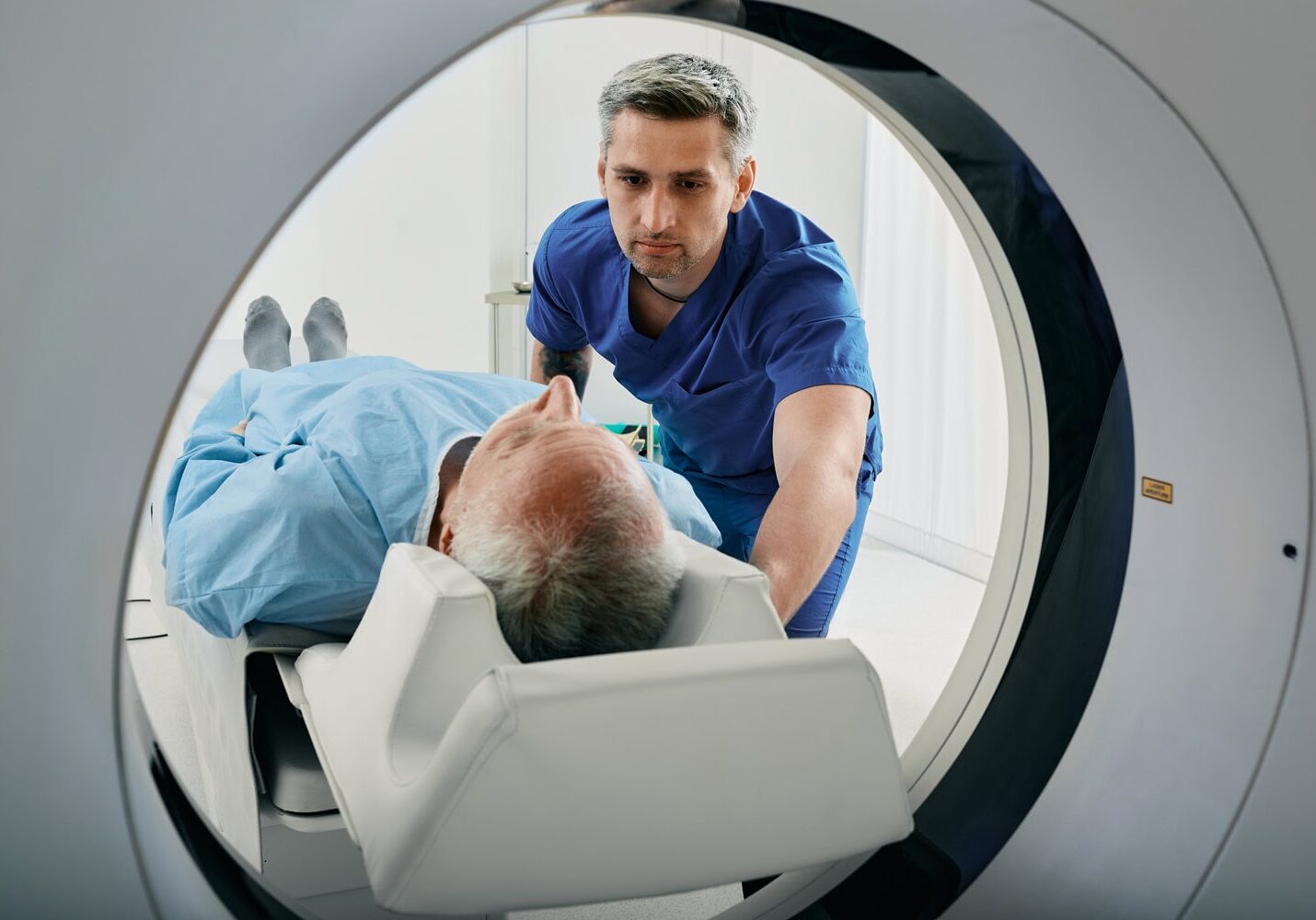 A healthcare professional in blue scrubs assists an older adult lying down for a scan inside an MRI machine.