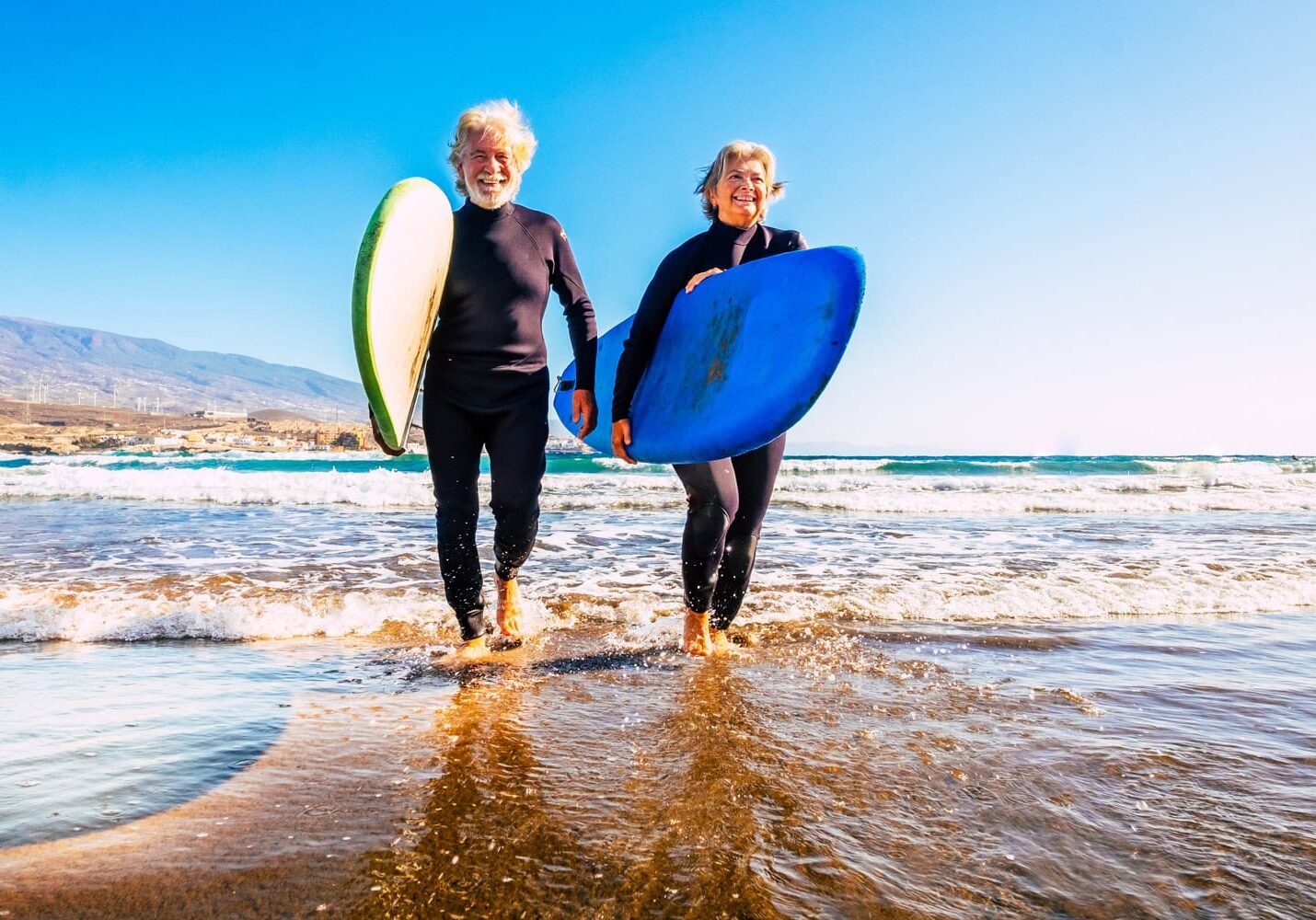 Two seniors in wetsuits carry surfboards while walking out of the ocean on a sunny day, showcasing that age is no barrier to adventure.
