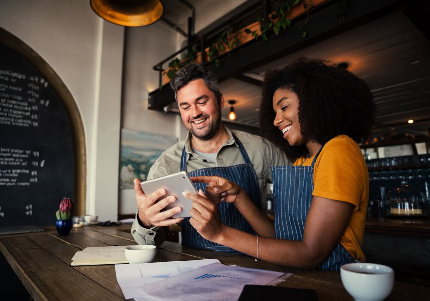Two people in aprons are smiling and looking at a tablet in a café, discussing their small business. A notebook, paper, and coffee cups are on the wooden table. One person is pointing at the tablet screen, likely reviewing SEO strategies to optimize their keywords for better online presence.