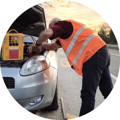 A person in a high-visibility vest works on the engine of a car with its hood open on the roadside, as if tinkering at home. A yellow toolbox is placed on the vehicle.