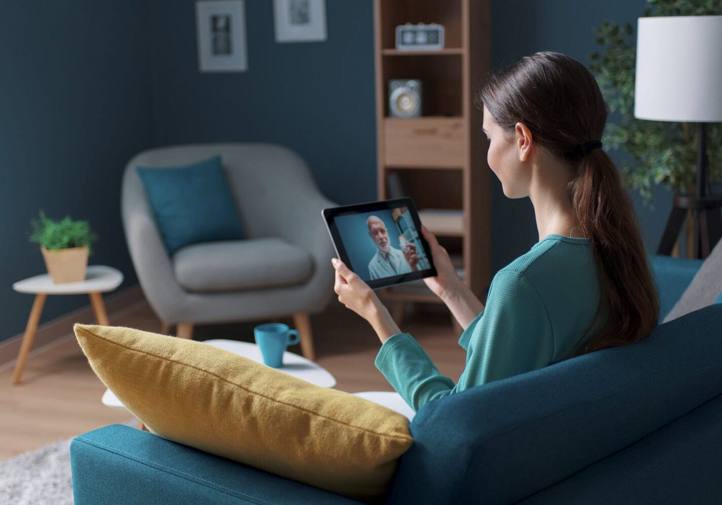 telemedicine-adult A woman sits on a blue sofa, holding a tablet and video chatting with a man. The room has modern decor, including a lamp, shelf, and an armchair with a plant on a side table. Their conversation revolves around freelance work opportunities for independent contractors.
