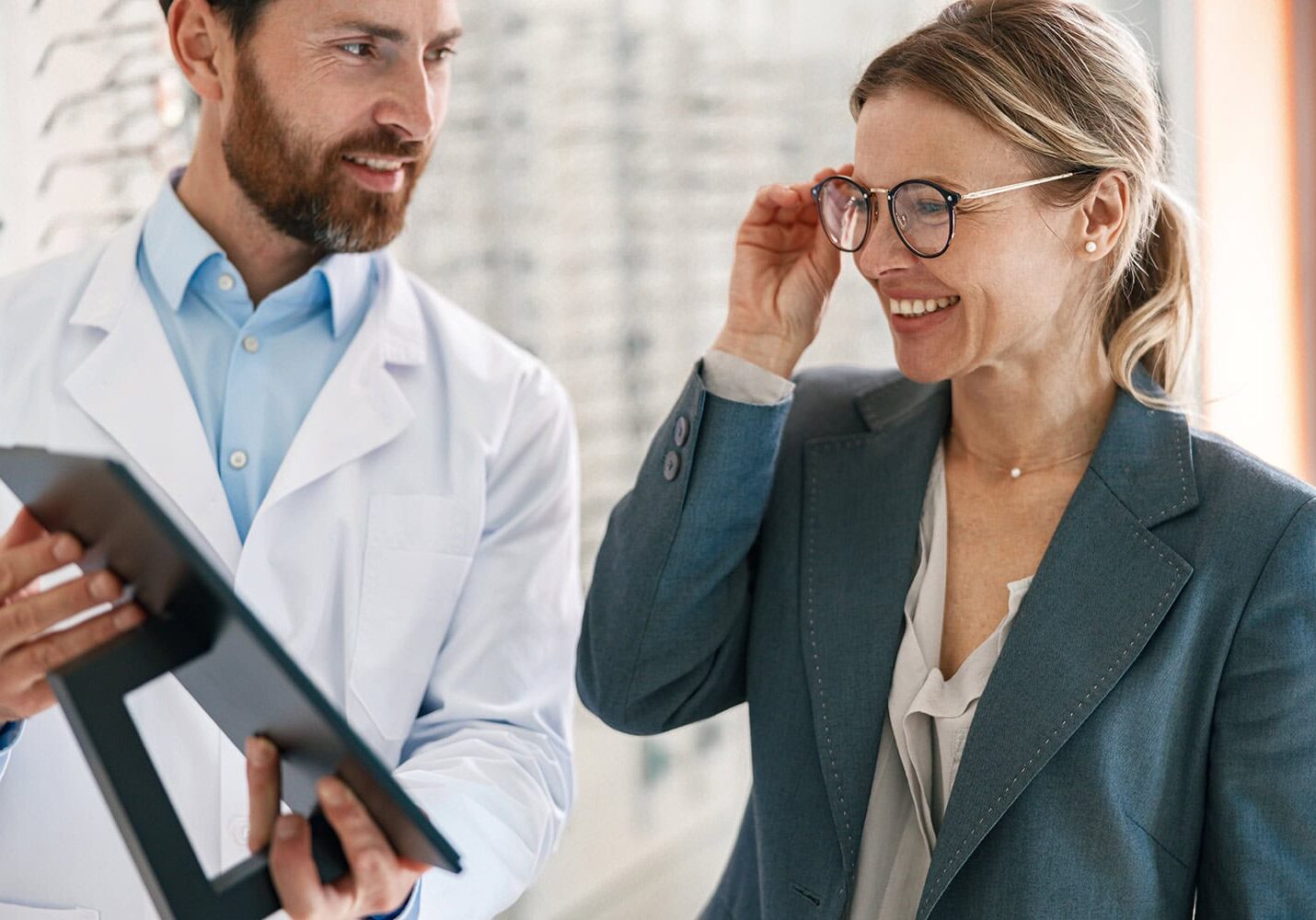 A man in a white coat holds a tablet and speaks with a smiling woman in glasses and a blazer, surrounded by groups of colleagues engaged in discussions.