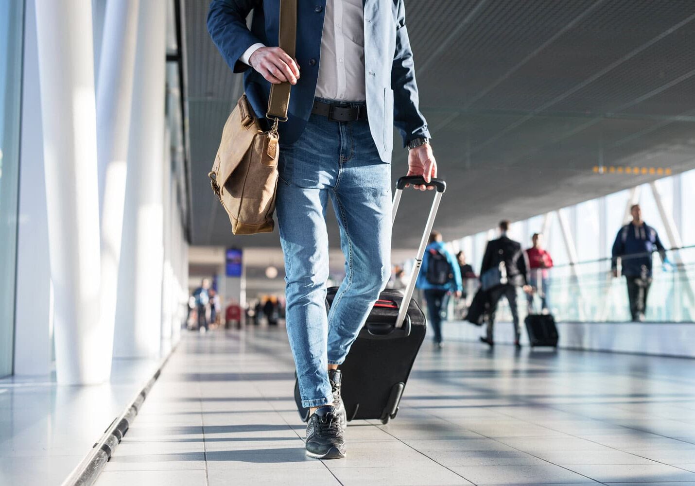 work-travel A person in jeans and a blazer walks through an airport terminal pulling a wheeled suitcase and carrying a shoulder bag, exuding the determination of a small business owner on the go.
