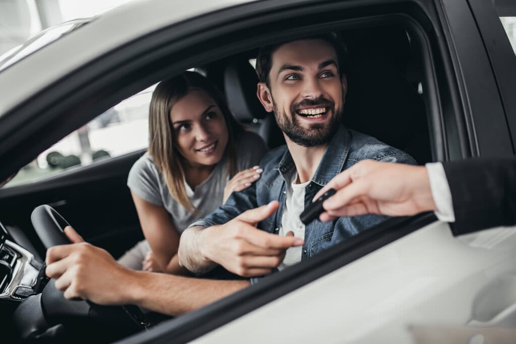 A smiling man in the driver’s seat of a car is receiving keys from a person outside the vehicle. A woman sits beside him, looking at the keys.