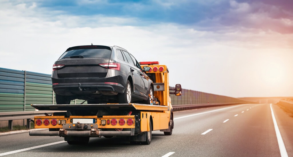 A black SUV, bearing the sleek emblem of Alliance Travel, is being transported on a flatbed tow truck down an empty highway.