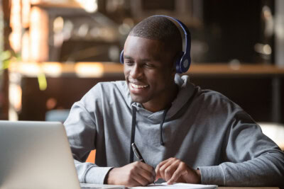 A person wearing headphones is seated at a table using a laptop while writing in a notebook, diligently preparing their college admission essay to apply to their dream school.
