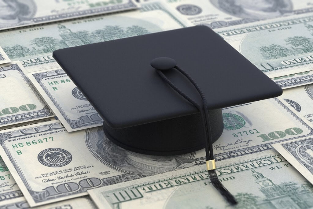 A graduation cap sits atop a spread of one hundred dollar bills, symbolizing the burden of college costs.