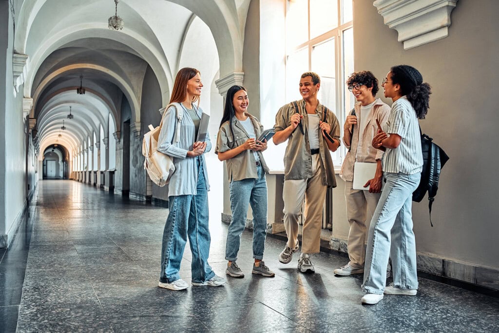 Five students with backpacks stand and talk in a hallway with arched ceilings and large windows, appearing relaxed and engaged in conversation about the Alliance College Guide.