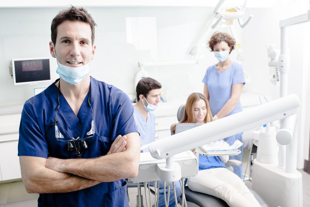 Alliance Health 2 A dentist in blue scrubs stands with arms crossed in a dental clinic, exemplifying professional dental care. In the background, two assistants and a seated patient can be seen.