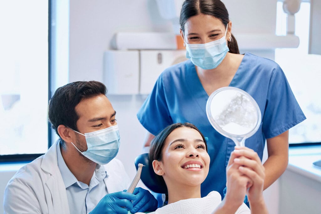 A dentist and dental assistant, both wearing masks, attend to a smiling patient in a dental chair who is holding up a mirror and looking at her teeth. The trio forms a cheerful group focused on maintaining dental health.