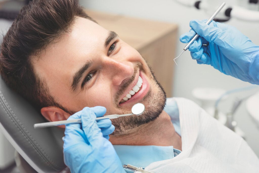 A man sits in a dental chair, smiling as a dentist wearing blue gloves holds dental instruments near his mouth during an examination, showcasing the exceptional service that highlights effective small business marketing.