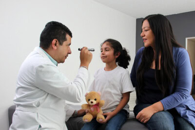 A doctor examines a young girl using an otoscope while she holds a teddy bear, illustrating the direct benefits of the healthcare alliance. A woman sits beside them, observing the interaction.
