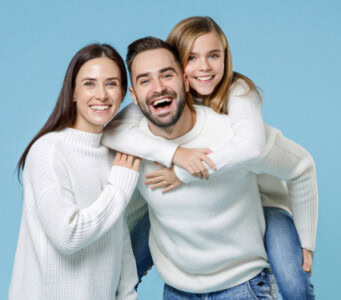 A man, woman, and young girl in white sweaters and jeans smile; the girl is giving the man a piggyback ride against a blue background, capturing the warmth of home.