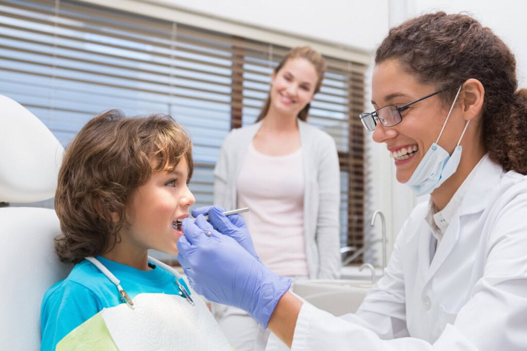 A dentist examines a young girl's teeth in a dental clinic while a woman, likely her mother, stands in the background watching and smiling, showcasing the importance of dental care for families.