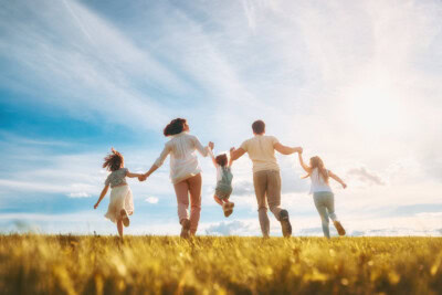A family of five, consisting of two adults and three children, is running hand-in-hand across a grassy field under a partly cloudy sky. The sun is shining brightly in the background, showcasing the joy and togetherness that highlight family benefits.