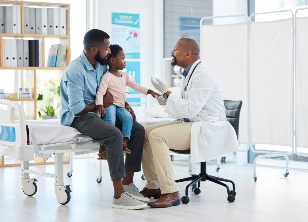 A doctor sits on a chair, engaging with a young girl sitting on her father's lap in a medical examination room, fostering an atmosphere where families feel cared for and supported.