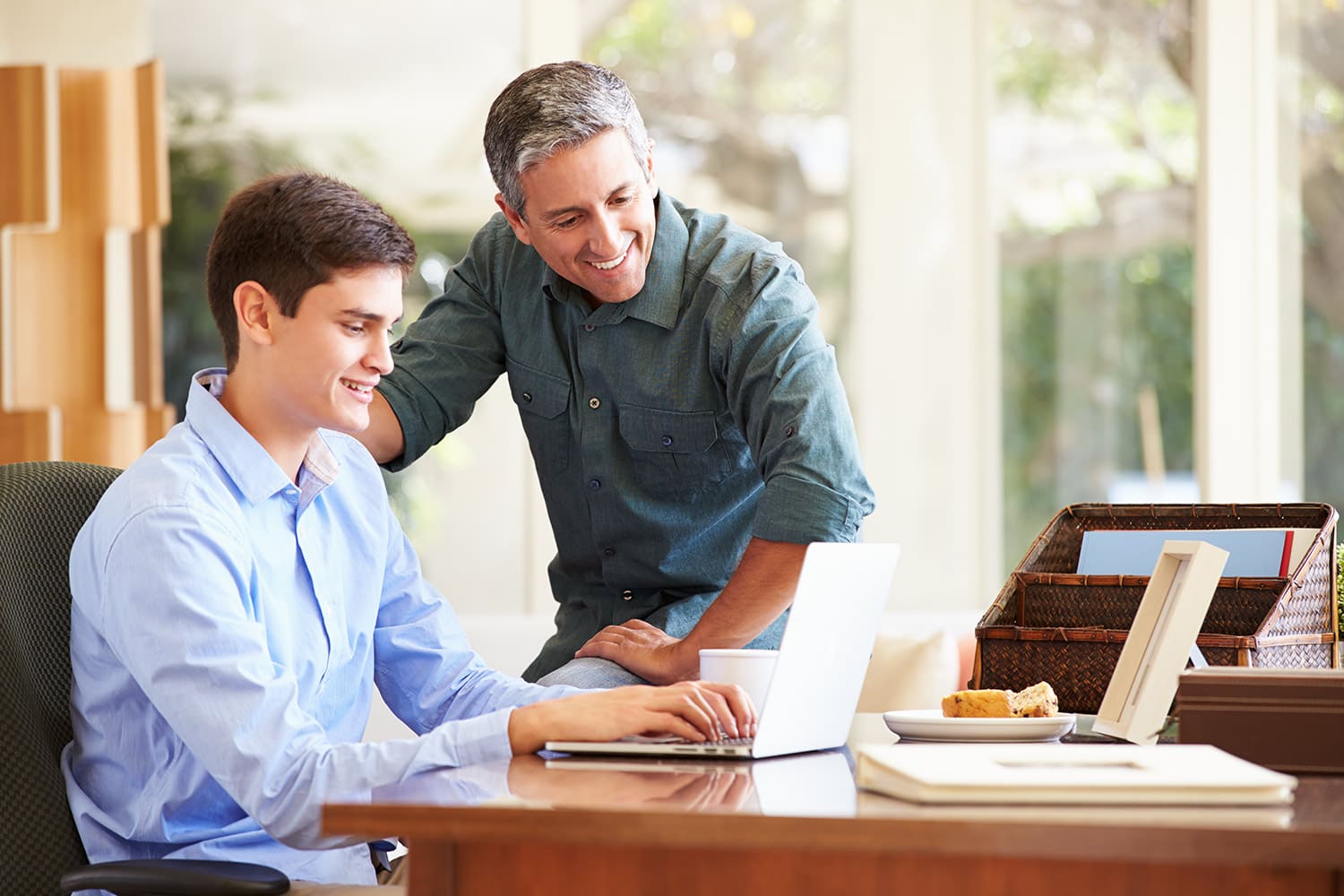 A man sits at a desk using a laptop while another man stands beside him, smiling and looking at the laptop screen. They are in a well-lit room with a window in the background, perhaps discussing how to prepare your child for college starting now with the help of a parent checklist.