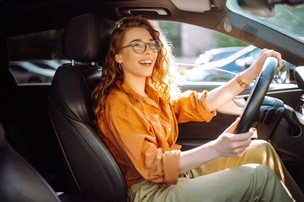 A woman with curly hair and glasses is sitting in the driver's seat of a car, smiling and holding the steering wheel. She is wearing an orange shirt and beige pants, embodying the freedom that comes with freelance work.