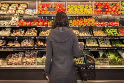 Person in a gray coat holding a basket and standing in front of a store produce section with various vegetables displayed, including bell peppers, mushrooms, and greens, carefully selecting their groceries amid rising prices.