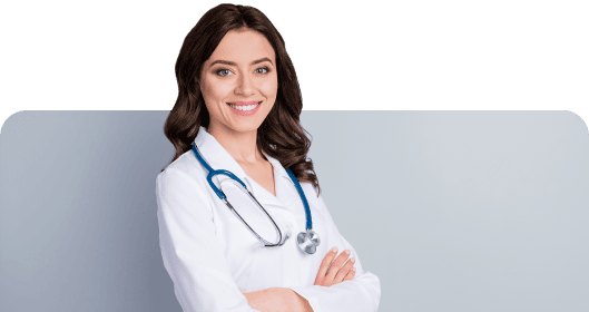 A smiling woman with long dark hair, wearing a white coat and a stethoscope around her neck, stands with arms crossed against a light gray background, embodying the trusted care you'd expect at home.
