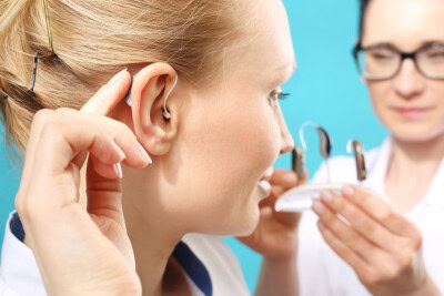 A person with blonde hair is adjusting a hearing aid in their ear while another, in the background, holds two hearing aids on a tray against a blue backdrop. The scene subtly emphasizes the importance of an EPIC service plan for optimal hearing solutions.